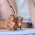 A hand holding a beige and silver pen, poised above a notepad, ready for writing. The pen features a logo.