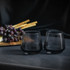A set of two smoked black whiskey glasses on a grey surface, with grapes and bamboo sticks in the background.