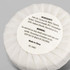 A white circular soap wrapped in paper, labelled with ingredients and warnings, features a logo.