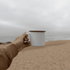 A drink bottle featuring a white enamel metal cup with a black rim, held against a beach backdrop.