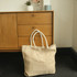 A herringbone patterned jute tote bag in natural and cream colours, with sturdy white handles, placed beside a wooden cabinet.