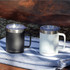 A drink bottle set featuring two Arctic Zone Titan Thermal Copper Mugs in black and white, each with a lid, on a striped cloth.