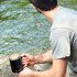 A drink mug in black with a stainless steel rim, held by a person by a riverside.