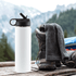A drink bottle in white with a black lid, placed beside hiking boots and a grey scarf against a mountain backdrop.