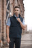 A men's black vest is featured, designed for workwear, with a zip closure and a logo. The model is posing outdoors.