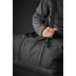 A black duffle bag is being held by a person, showcasing its sturdy handle and sleek design. The bag has a logo.