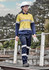 A woman in a yellow and navy lightweight ripstop taped shirt walks on a wooden path, wearing a helmet and holding gloves.