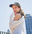 A woman in a black cap and white shirt poses outdoors, with a city skyline in the background. The cap features a logo.