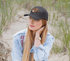 A black premium American twill cap with a logo, worn by a person with long hair, set against a sandy background.