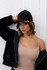 A black cap with flame graphics is worn by a model in a beige top and black jacket, posed against a white background.