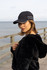 A black microfibre sports cap with white piping, worn by a woman on a seaside boardwalk.