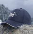 A navy sports cap with sandwich trim, featuring a logo on the front, resting on a stone in a mountainous landscape.