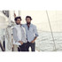 Two men posing on a sailboat, one wearing a light blue button-up shirt and the other in a white vest over a checked shirt.