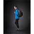 A woman in a blue lightweight softshell jacket, wearing black leggings and a backpack, standing against a dark background.