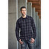 A man wearing a black and grey checked button-up shirt, standing against a concrete wall. The shirt has a logo.