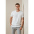 A man in a white crew neck t-shirt leans against a wall, smiling and wearing light grey pants.