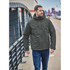 A man wearing a dark green thermal shell jacket, walking outdoors along a paved path. The jacket features a hood.