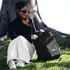 A black Thule Recycled Lumion 16" computer backpack sits on the grass beside a person wearing sunglasses.