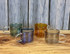 Four transparent double-wall mugs in green, amber, blue, and grey arranged on a wooden surface against a textured background.