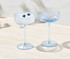 Two clear blue coupe glasses, one filled with ice and blueberries, elegantly presented on a light surface.