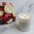 A large white glass candle with a lit wick beside a bouquet of cream and pink flowers.