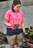 A woman in a pink and navy polo shirt holds flowers, wearing khaki cargo shorts, surrounded by gardening supplies.