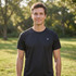 A black T-shirt featuring a logo, worn by a man standing in a grassy outdoor setting with trees in the background.