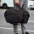 A black duffle bag is being held by a person, featuring dual handles and a shoulder strap.