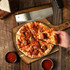 A wooden pizza board with a pizza topped with pepperoni and cheese, alongside dipping sauce and a knife.