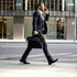 A man in a suit carries a black laptop conference bag while walking down a city street. The bag has a logo.