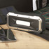 A waterproof outdoor Bluetooth speaker in black and silver, placed next to a smartphone on a wooden surface.
