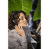 A woman with curly hair wears black headphones while enjoying music. Green foliage is visible in the background.