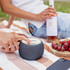 A circular, grey wireless speaker with a wooden top sits on a picnic blanket, alongside a bottle of wine and grapes.