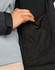 A close-up of a hand reaching into a black pocket on a softshell jacket. The jacket features a contrasting grey layer.