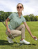 A light green polo shirt is worn by a woman crouching on grass, holding a golf ball, with sunglasses and casual trousers.