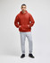 A man wearing a red pullover hooded sweatshirt and grey sweatpants, standing against a plain background.