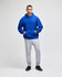 A blue pullover hooded sweatshirt with a front pocket, paired with grey sweatpants. The model stands confidently.
