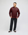 A man stands wearing a burgundy hooded sweatshirt and black pants. The sweatshirt features a logo on the sleeve.