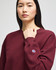 A burgundy women's crewneck sweatshirt featuring a logo on the sleeve. The model is standing and looking forward.