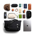 A black messenger bag with various items including sunglasses, a hat, a laptop, and a wallet, arranged on a flat surface.
