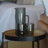 A drink bottle and two tumblers in smoky grey, placed on a round table beside a couch. The items feature a sleek design.