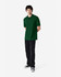 A man stands wearing a green Gildan Softstyle adult double pique polo, paired with black pants and shoes.