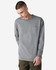 A man is wearing a grey long-sleeve t-shirt with a pocket, standing in a neutral backdrop. The shirt has a logo.
