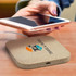 A square wireless charger in a beige hue, with a logo, being used with a smartphone on a wooden surface.
