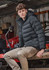 A unisex hooded puffer jacket in black, featuring a fitted design and a logo, worn by a model sitting on machinery.