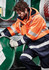 A man in an orange flame-resistant jacket adjusts a green barrel, wearing gloves and dark trousers.