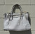 Soft, buttery leather meets a structured silhouette with polished gold-tone hardware and a clean, crisp white finish. Featuring dual rolled handles plus a detachable, adjustable crossbody strap, this satchel transitions effortlessly from work to weekend. Roomy interior with slip and zip pockets keeps essentials organized on the go.