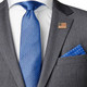 The gold rectangle American flag pinned to the right lapel of a gray suit jacket in a torso-only view and paired with a sky blue tie and white dress shirt.