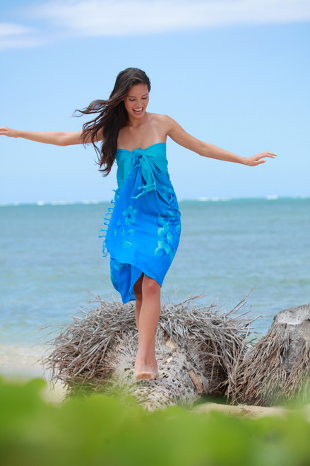 Woman wearing Blue Aloha Sarong  & walking on fallen tree on a sandy beach with water in the background
