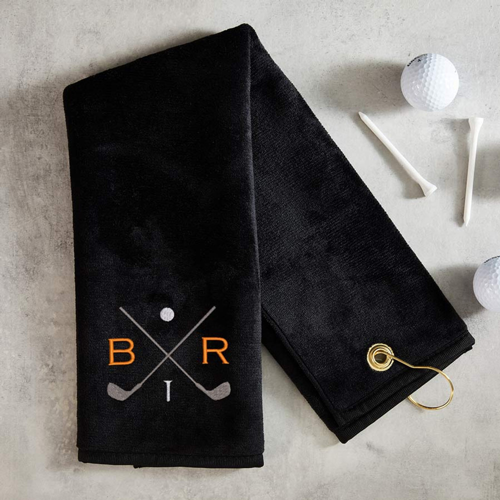 Custom Monogrammed Golf Towel in black embroidered with crossed golf clubs in gray thread, a golf ball and a golf tee in white thread, and your first and last initial in the thread color of choice, shown folded on table with golf balls and tees.